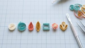 Row of basic paper quilling shapes on a cutting mat: tight coil, loose coil, teardrop, marquise, square, fringed flower and husked petal, beside a slotted quilling tool and tweezers
