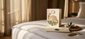 Handmade Vietnamese quilling card with a raised lotus design and blank logo medallion placed on a hotel bed at turndown beside a wax-sealed envelope and jasmine sprig, illustrating corporate and hospitality quilling card gifting