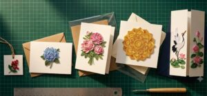 Top-down flat lay of handmade Vietnamese quilling cards in five formats, from a mini gift tag and flat postcard to a 5x7 folded card, a square mandala card and a tall gatefold crane card, with kraft, cream and navy envelopes, clear sleeves and a steel ruler on a green cutting mat