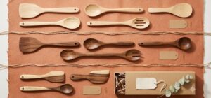 Overhead flat lay of Vietnamese wooden kitchen utensils arranged in three tiers on terracotta paper: pale rubberwood basics, richer acacia pieces and premium two tone acacia items beside a kraft gift box, illustrating a good, better, best wooden kitchenware range